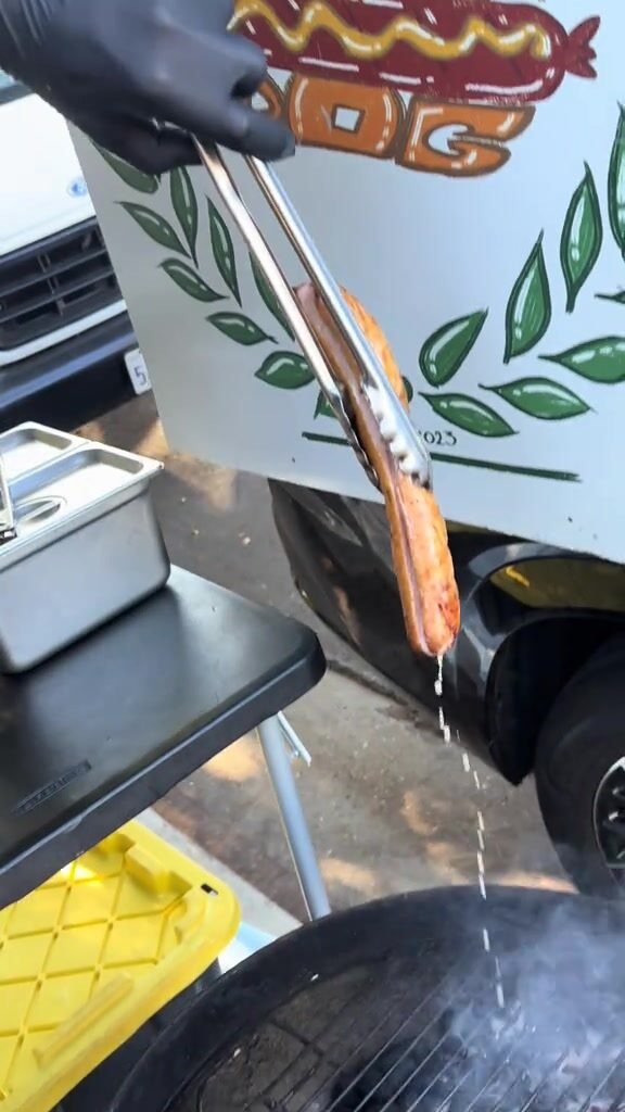 A sausage lifted off the grill with tongs in front of the Special Hot Dog banner