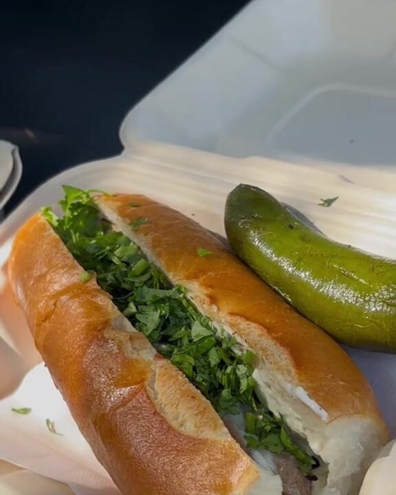 A finished hot dog in a toasted bun with fresh herbs and a pickled pepper on the side