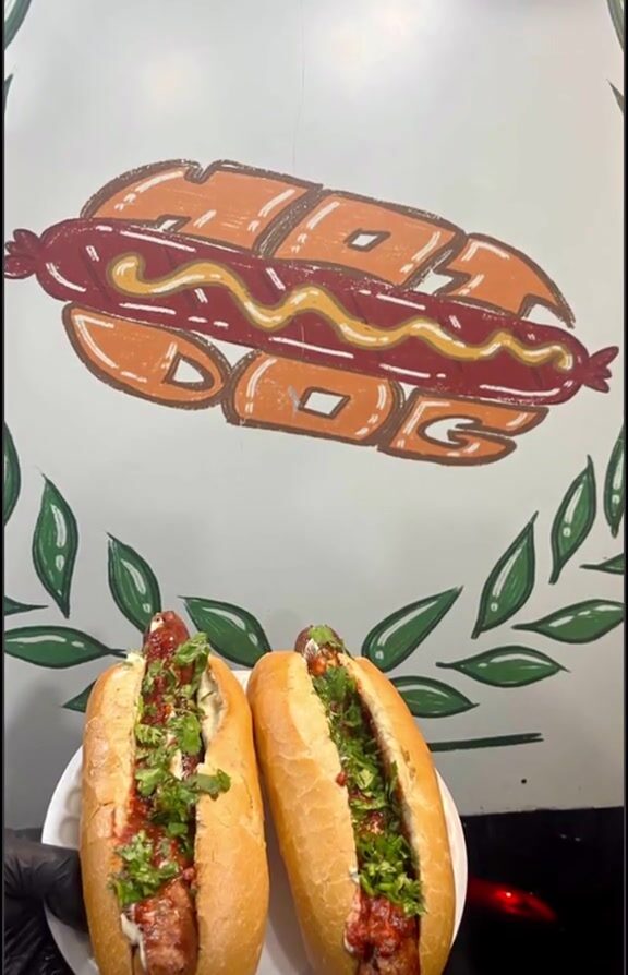Two Special Hot Dogs dressed with herbs and red sauce, held up in front of the Special Hot Dog banner